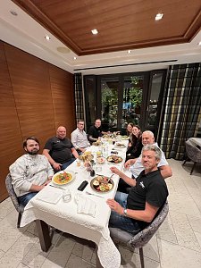 Acht Personen sitzen gemeinsam an einem Tisch in einem eleganten Restaurant mit Holzvert&auml;felung und Blick ins Gr&uuml;ne. Vor ihnen stehen Teller mit warmen Speisen. Die Gruppe blickt l&auml;chelnd direkt in die Kamera.