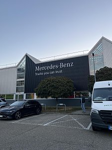 Ein modernes Geb&auml;ude mit einer dunklen Fassade tr&auml;gt den gro&szlig;en wei&szlig;en Schriftzug Mercedes-Benz Trucks you can trust. Davor erstreckt sich ein gepflasterter Parkplatz.