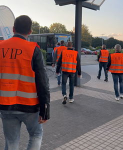 Mehrere Personen von hinten gesehen gehen in leuchtend orangefarbenen VIP-Warnwesten &uuml;ber ein Au&szlig;engel&auml;nde auf einen bereitstehenden Bus zu.