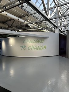 Eine geschwungene, wei&szlig;e Wand steht in einer modernen Halle mit gl&auml;nzendem, grauem Boden und einer offenen, verglasten Dachkonstruktion aus Stahltr&auml;gern. Auf der Wand prangt der hellgr&uuml;ne Schriftzug CHARGED TO CHANGE.