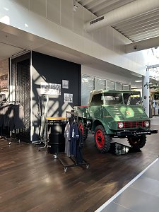 Ein historischer, gr&uuml;ner Unimog mit roten Felgen steht als Ausstellungsst&uuml;ck auf einem dunklen Holzboden. Links daneben sind Kleiderst&auml;nder mit Merchandise-Artikeln und dekorative schwarze F&auml;sser platziert.