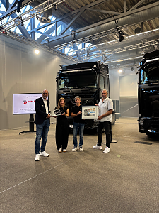 Vier Personen stehen nebeneinander f&uuml;r ein &Uuml;bergabefoto vor einem gro&szlig;en, schwarzen Mercedes-Benz Lkw in einer hellen Werkshalle. Sie halten ein gerahmtes Bild sowie ein gro&szlig;en Schl&uuml;ssel in der Hand.