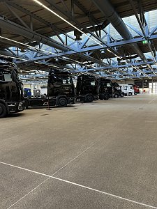 Mehrere schwarze Mercedes-Benz Lkw-Zugmaschinen stehen schr&auml;g hintereinander aufgereiht in einer weiten Produktionshalle. Der Blickwinkel zeigt die Fahrzeuge von der Seite auf einer grauen Betonfl&auml;che.