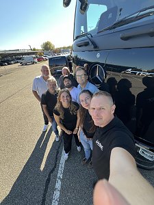 Eine Gruppe von acht Personen posiert f&uuml;r ein Selfie direkt neben der gl&auml;nzenden, schwarzen Front eines Mercedes-Benz Lkw. 