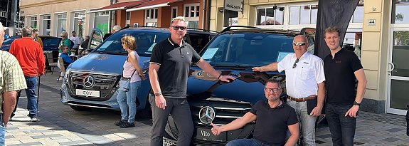 Vier M&auml;nner posieren vor einem schwarzen Mercedes Vito auf einer Kopfsteinpflasterstra&szlig;e in Heiligenstadt. Links und rechts im Hintergrund stehen weitere Fahrzeuge, im Hintergrund sind mehrst&ouml;ckige Altstadth&auml;user mit Schaufenstern zu sehen. 