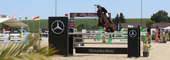 Das Bild zeigt einen Springreiter in Immenrode, der mit seinem Pferd &uuml;ber ein Hindernis mit Mercedes-Benz-Werbung springt. Im Hintergrund sieht man eine Reitturnier-Arena mit verschiedenen Hindernissen, Fahnen mehrerer L&auml;nder und Zuschauerbereichen. Das Wetter ist sonnig, und es gibt einige Pflanzen und Dekorationen um die Arena.
