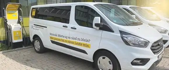 Weißer Miet-Van mit ADAC-Beschriftung steht an einer Ladesäule; seitlicher Schriftzug: „Normale Mietwagen sind zu klein? Hier passen bis zu neun Leute rein.“