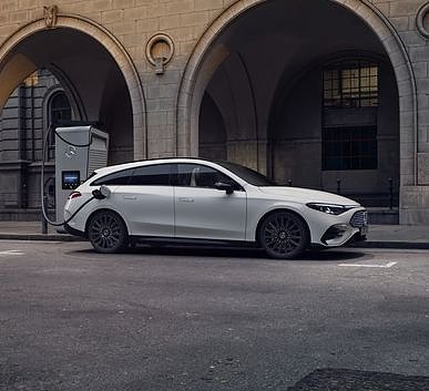 Das Bild zeigt einen weißen Mercedes-Benz CLA Shooting Brake neben einer Ladesäule, der auf einer breiten, nahezu leeren Stadtstraße steht. Moderne Gebäude umgeben die Szene, das Licht wirkt weich und frühmorgendlich. Der Wagen ist seitlich zu sehen und vermittelt Dynamik sowie Eleganz.