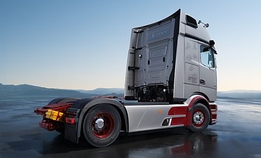 Graues Lkw-Fahrerhaus von hinten rechts. Zwei Achsen mit roten Felgen. Silberne Verkleidung an der Seite mit roten Akzenten. Der Lkw steht auf nassem, spiegelndem Asphalt vor Bergen und hellblauem Himmel.