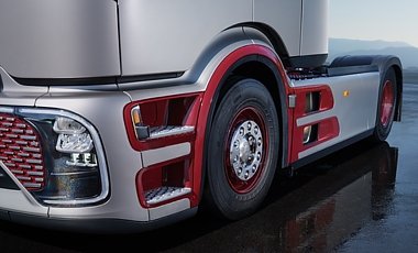 Nahaufnahme der Lkw-Front und Seitenverkleidung. Silberner Lack mit auff&auml;lligen, roten Umrandungen an den Einstiegsstufen und Reifen-Radl&auml;ufen. Gro&szlig;er Scheinwerfer links. Vorderrad mit roter Felge auf nassem Asphalt