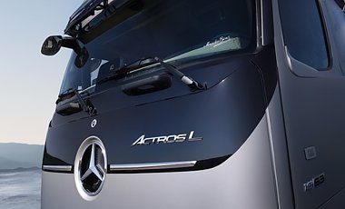 Nahaufnahme der Lkw-Front. Unten silberner Lack, dar&uuml;ber eine gl&auml;nzende, schwarze Fl&auml;che. Links prallt das verchromte Mercedes-Benz-Logo hervor. Rechts daneben steht der geschwungene Schriftzug Actros L in Silber.