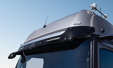 Dachpartie eines silbernen Lkws von vorne rechts. &Uuml;ber der Windschutzscheibe ist eine dunkle Sonnenblende mit integrierten LED-Leuchten angebracht. Oben auf dem Dach sind Antennen zu sehen. Blauer Himmel.