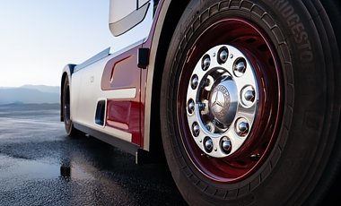 Nahaufnahme eines Lkw-Vorderrads. Gro&szlig;er, schwarzer Reifen mit dunkelroter Felge. In der Mitte gl&auml;nzt eine verchromte Radkappe mit Mercedes-Stern. Im Hintergrund ist die silber-rote Seitenverkleidung auf nassem Asphalt zu sehen.