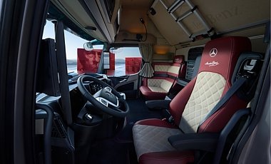 Moderner Lkw-Innenraum von vorne links fotografiert. Schwarzes Lenkrad mit Kn&ouml;pfen im Vordergrund. Beigefarbene Ledersitze mit roter Umrandung und Mercedes-Logo. Im Hintergrund eine gem&uuml;tliche Sitzecke. 