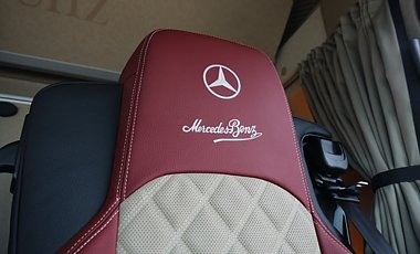 Kopfst&uuml;tze eines Lkw-Sitzes im Detail. Rotes Leder mit wei&szlig;er Ziernaht. Aufgestickt sind das runde Mercedes-Benz-Logo und darunter der geschwungene Schriftzug. Rechts im Hintergrund h&auml;ngt ein geraffter, grauer Vorhang vor dem Fenster.