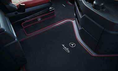 Schwarzer Lkw-Fu&szlig;boden im Detail. Die passgenauen Teppiche haben eine auff&auml;llige, rote Umrandung. Auf dem Teppich im Vordergrund sind das wei&szlig;e Mercedes-Benz-Logo und der geschwungene Markenschriftzug aufgestickt.