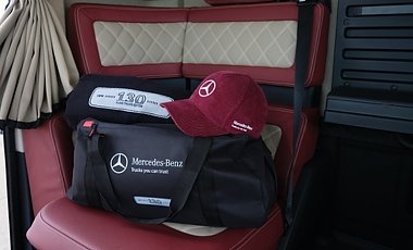 Fan-Artikel auf einem roten Lkw-Sitz. Eine gro&szlig;e, schwarze Reisetasche mit wei&szlig;em Mercedes-Benz Logo liegt auf der Sitzfl&auml;che. Darauf liegen eine dunkelrote Baseballkappe mit wei&szlig;em Stern und eine gefaltete, schwarze Decke mit der Zahl 130.